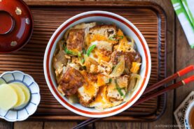 A bowl of Simmered Tofu and Egg Rice Bowl with tofu, softly cooked eggs, and onions simmered in a savory sauce, then spooned over steamed rice&mdash;placed on a wooden tray with pickled ginger, chopsticks, and a miso soup bowl nearby.