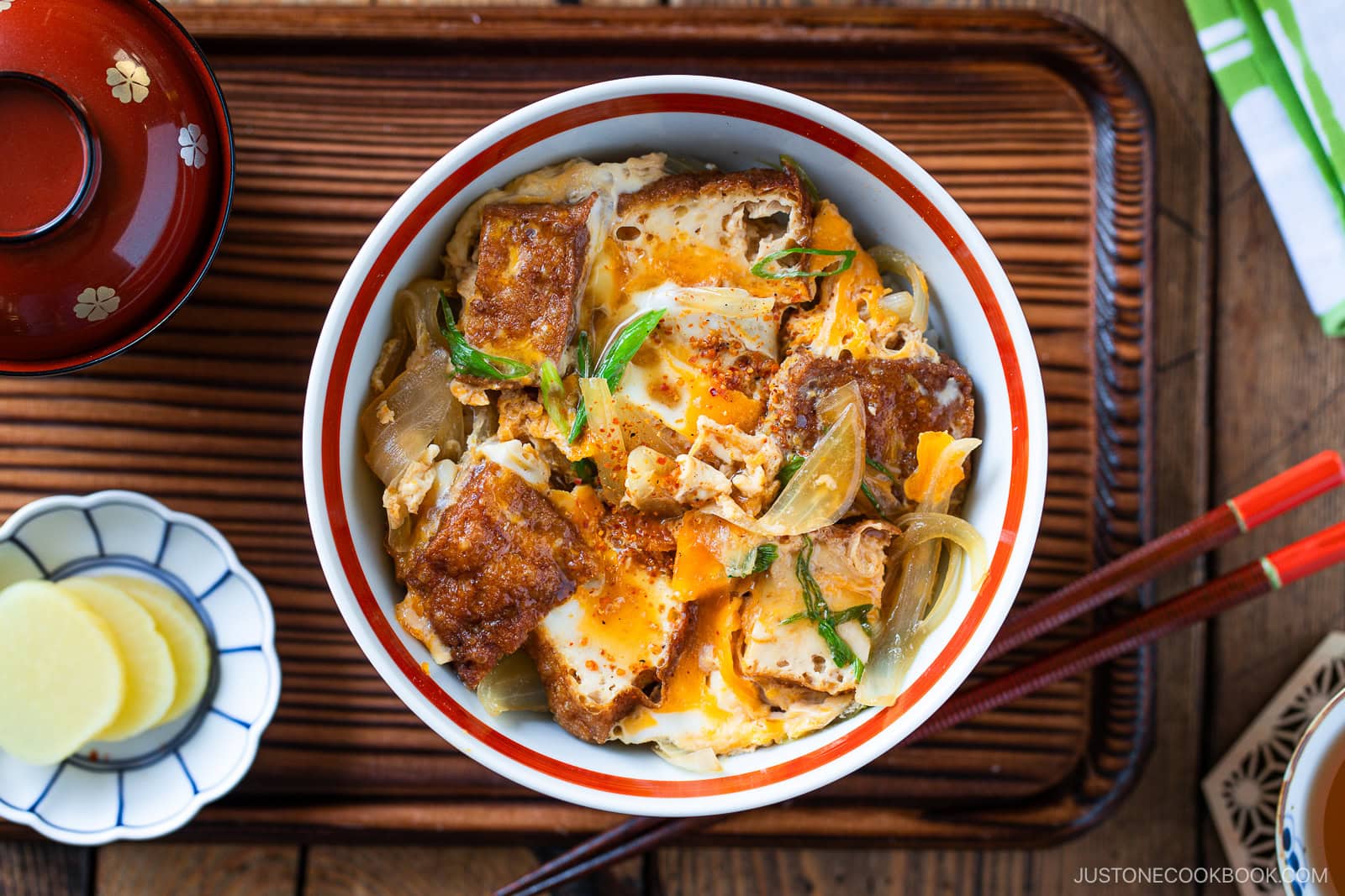 A bowl of Simmered Tofu and Egg Rice Bowl with tofu, softly cooked eggs, and onions simmered in a savory sauce, then spooned over steamed rice&mdash;placed on a wooden tray with pickled ginger, chopsticks, and a miso soup bowl nearby.