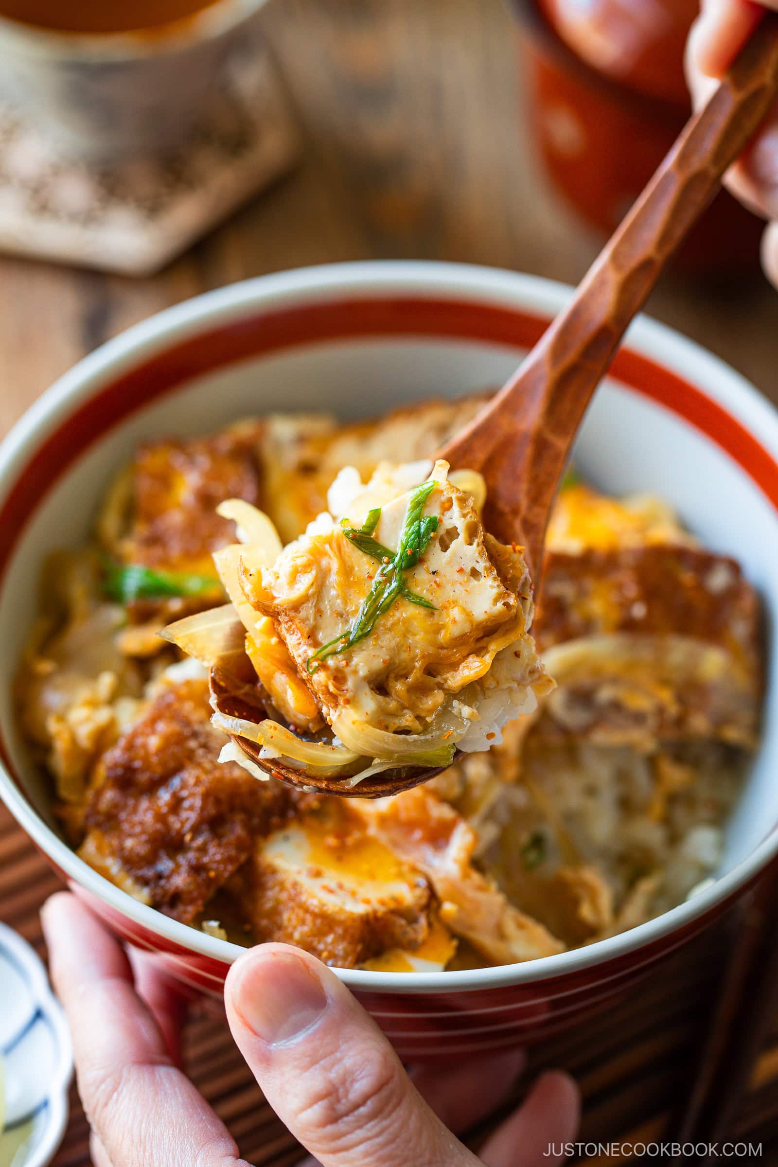 A close-up of a hand holding a wooden spoon with a bite of Simmered Tofu and Egg Rice Bowl&mdash;fried tofu, egg, and onions&mdash;a reminiscent of comforting classics like oyakodon.