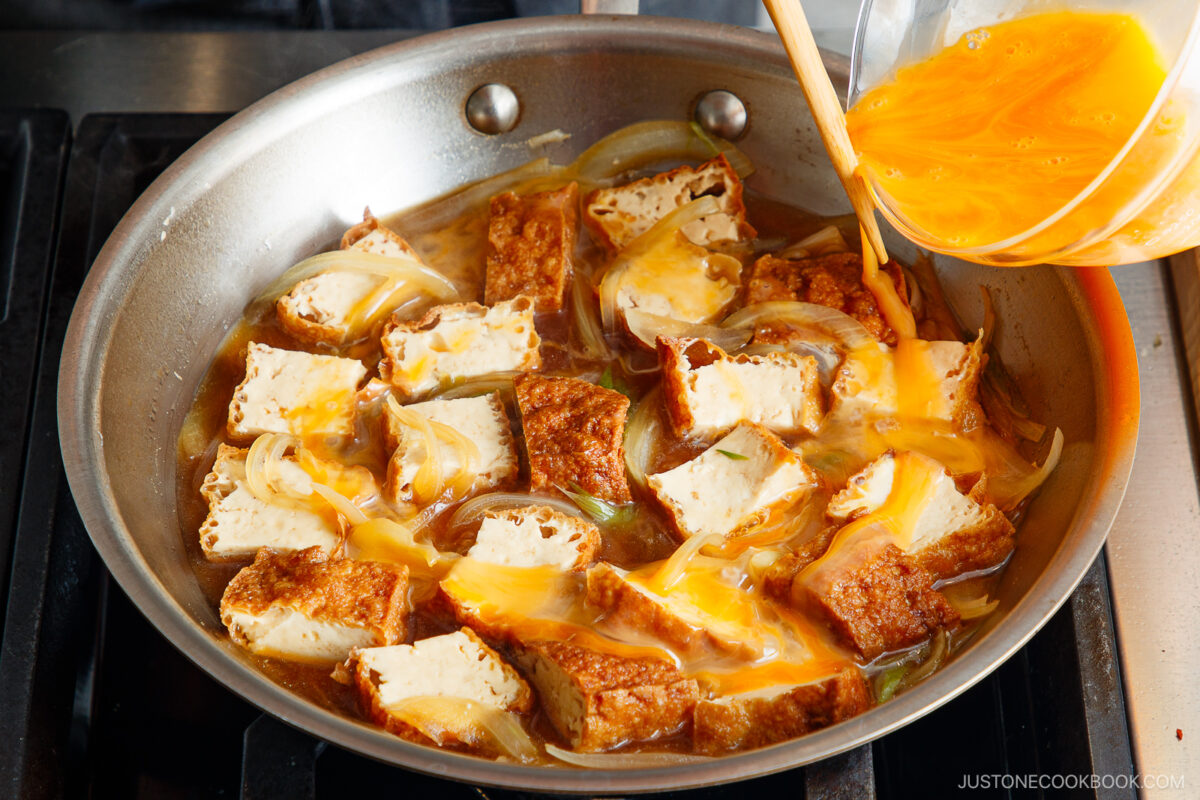 A pan filled with browned tofu and sliced onions on a stovetop, while beaten eggs are being poured over the ingredients from a glass bowl, ready to cook this Simmered Tofu and Egg Rice Bowl.