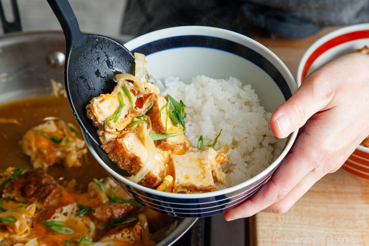 A hand holds a bowl of white rice topped with simmered tofu and onions, creating a delicious Simmered Tofu and Egg Rice Bowl, while a black spoon serves the tofu mixture from a pan in the background.