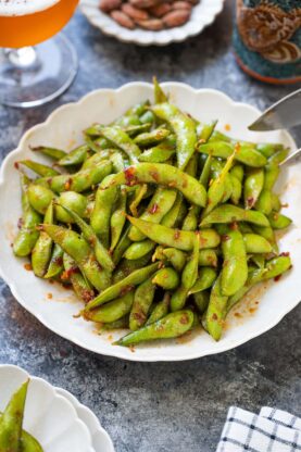 A white scalloped bowl filled with spicy edamame pods coated in a bold red seasoning, placed on a dark tabletop next to a drink and a small bowl of nuts.