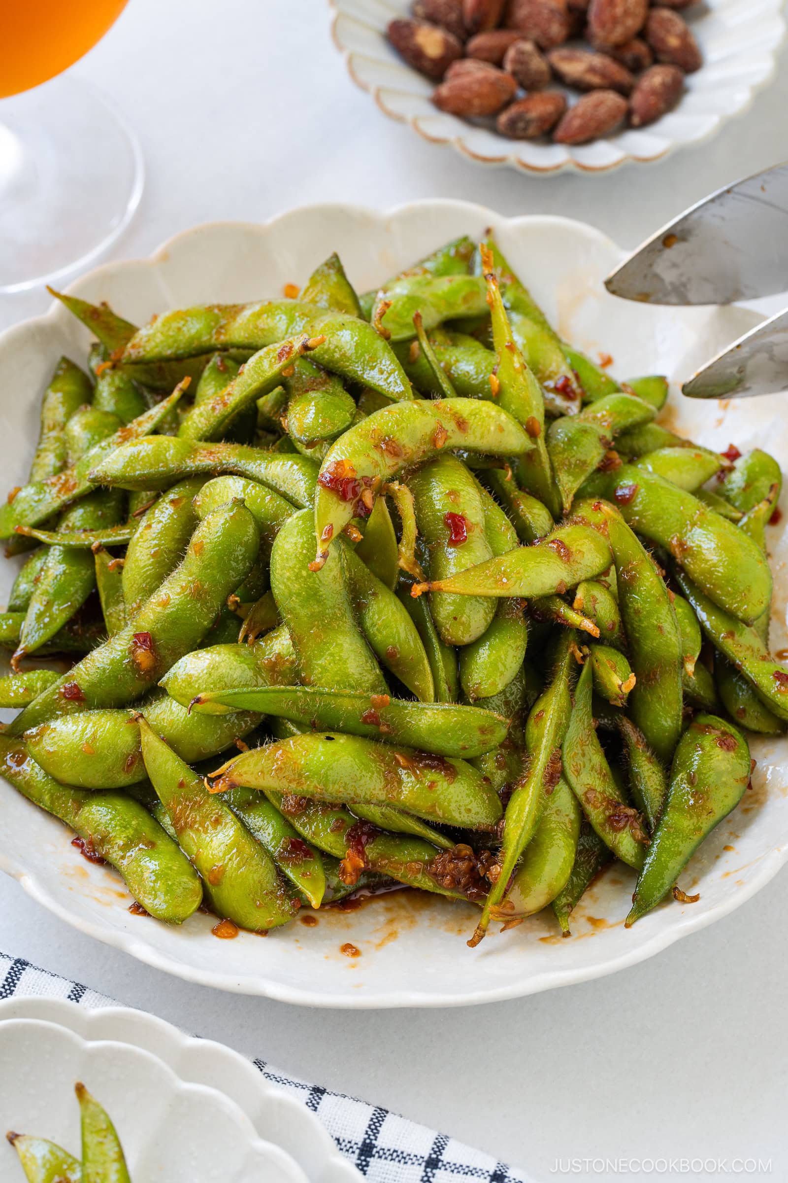 A white plate of spicy edamame, garnished with chili flakes, sits on a table next to a bowl of almonds and a glass of orange beverage.