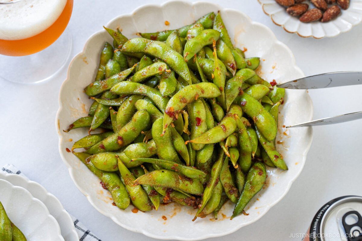 A white bowl filled with spicy edamame pods coated in savory spices, with metal tongs resting on the edge. Part of a glass of beer and a small plate of almonds are visible in the background on a white table.
