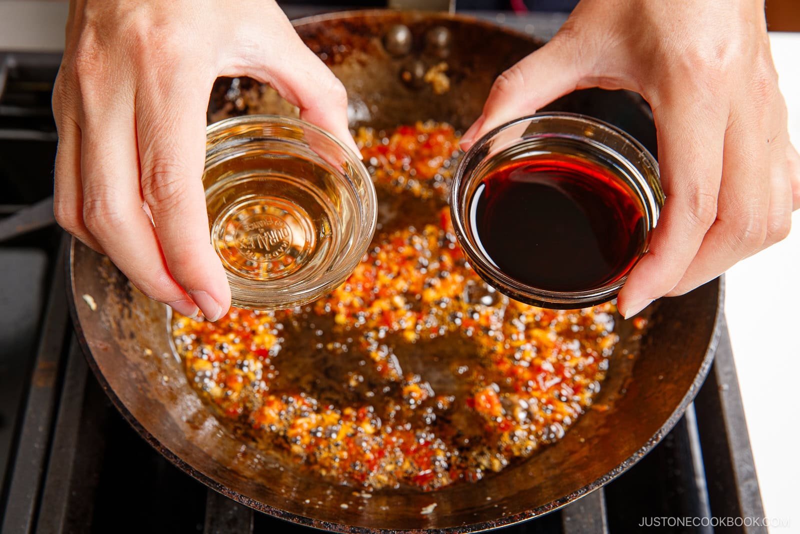Two hands hold small glass bowls, one with clear liquid and one with dark liquid, over a pan with sizzling chopped garlic, chilies, and oil—perfect for preparing spicy edamame on the stove.
