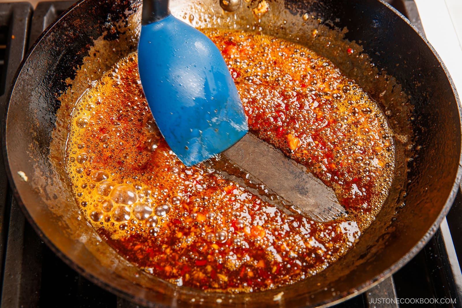 A blue spatula stirs a bubbling, reddish-brown spicy edamame sauce with oil and spices in a black frying pan on a stovetop.