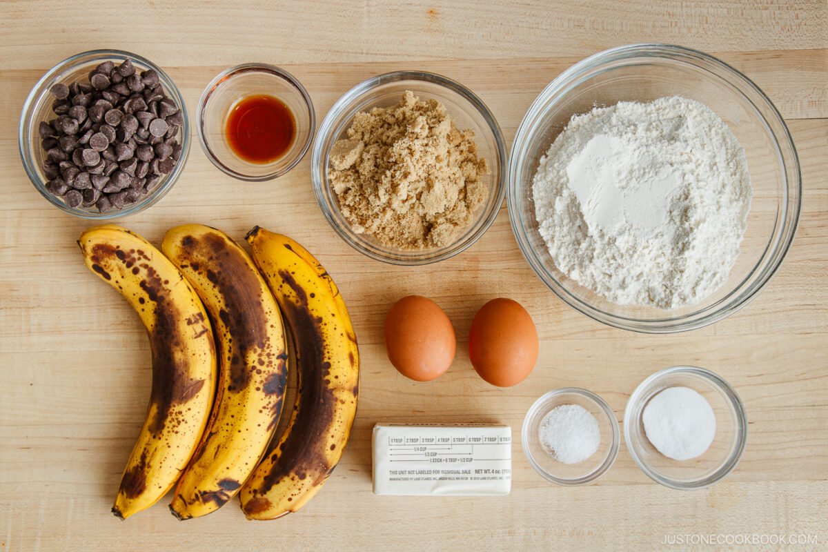 Top-down view of baking ingredients for Brown Butter Banana Bread on a wooden surface: three ripe bananas, two eggs, chocolate chips, vanilla extract, brown sugar, flour, browned butter, baking powder, and baking soda in separate bowls.