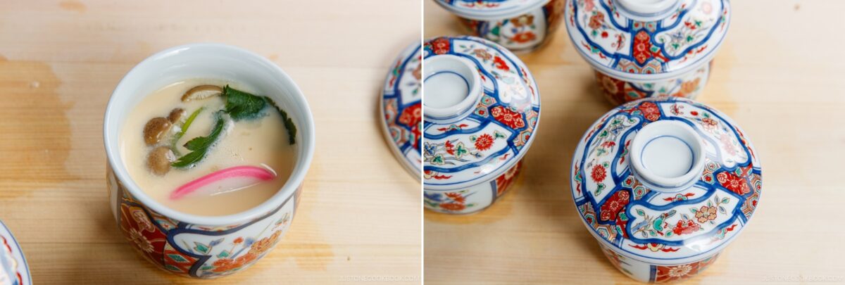 A close-up of a traditional Japanese cup containing Chawanmushi (Japanese Savory Steamed Egg Custard) with mushrooms, herbs, and a pink garnish, sits beside three matching lidded cups on a wooden surface.