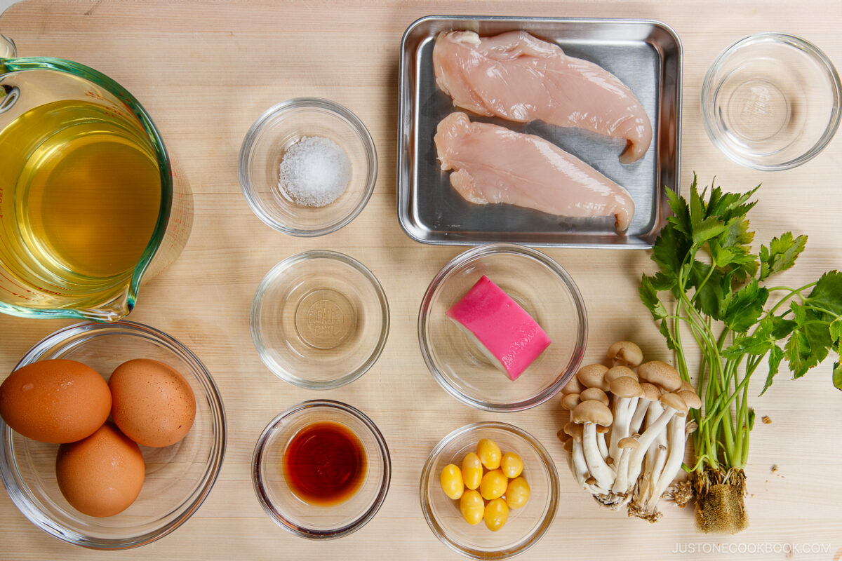 Top-down view of Chawanmushi (Japanese Savory Steamed Egg Custard) ingredients on a wooden surface, featuring chicken breasts, eggs, broth, mushrooms, fresh parsley, salt, soy sauce, gingko nuts, fish cake slice, and small glass bowls.