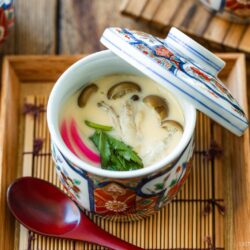 A colorful cup of Chawanmushi (Japanese Savory Steamed Egg Custard), garnished with mushrooms, herbs, and fish cake, sits on a wooden tray with a red spoon beside it. The lid is slightly open.