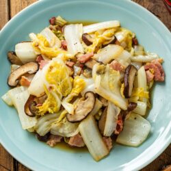 A blue plate filled with Napa Cabbage Stir-Fry—sliced mushrooms and bacon—rests on a wooden table, with part of a rice bowl and soup at the edges, inspired by Just One Cookbook.