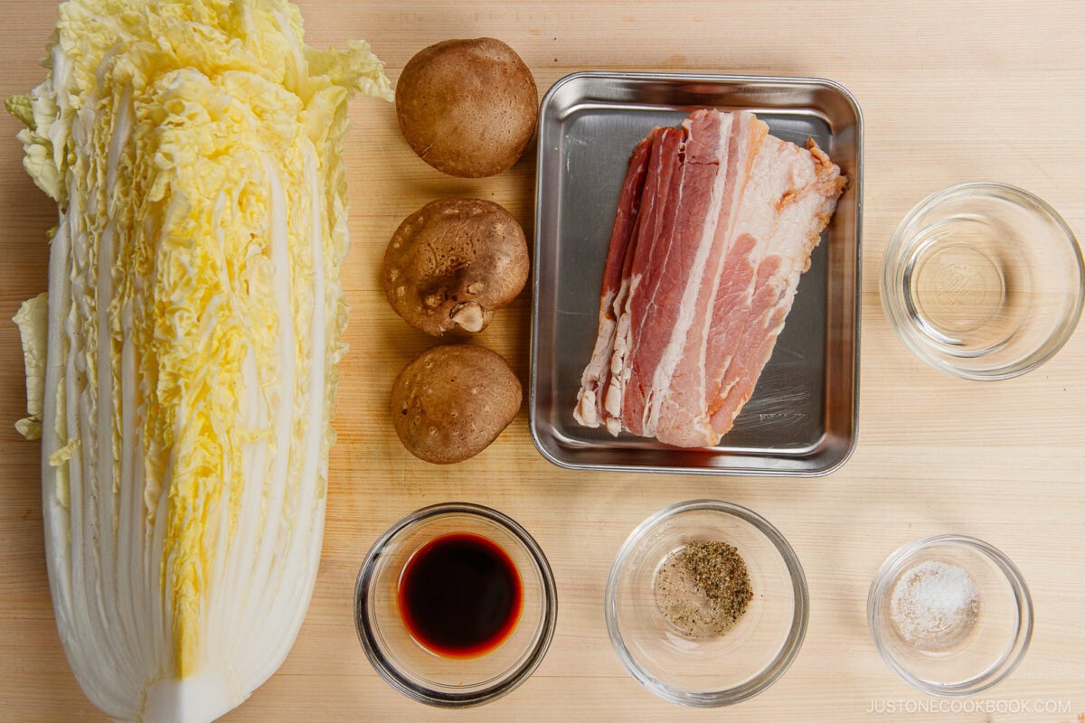 Top-down view of Napa cabbage, three mushrooms, sliced bacon, and seasoning bowls arranged neatly on wood—perfect ingredients for a delicious Napa Cabbage Stir-Fry inspired by Just One Cookbook.