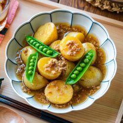 A bowl of Japanese-style Simmered Potato and Chicken with minced meat and green snap peas, served on a wooden tray with chopsticks, a drink with ice, and a bowl of rice nearby.