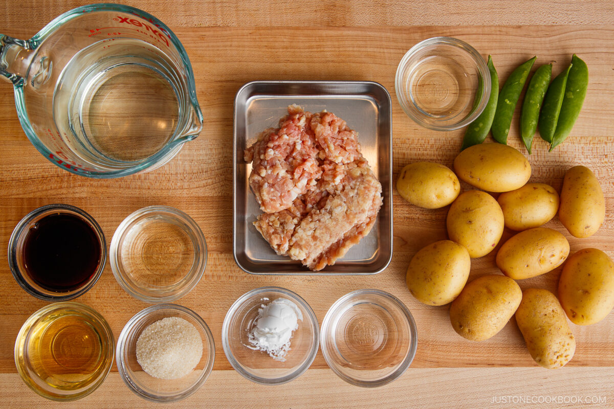 Overhead view of raw cooking ingredients for Simmered Potato and Chicken on a wooden surface: ground meat, baby potatoes, snow peas, clear measuring cup with liquid, sliced onions, and small bowls with soy sauce, sugar, and seasonings.
