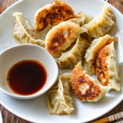 A white plate with golden-brown pan-fried dumplings, made from a classic gyoza recipe, arranged around a small bowl of soy dipping sauce, set on a wooden table.