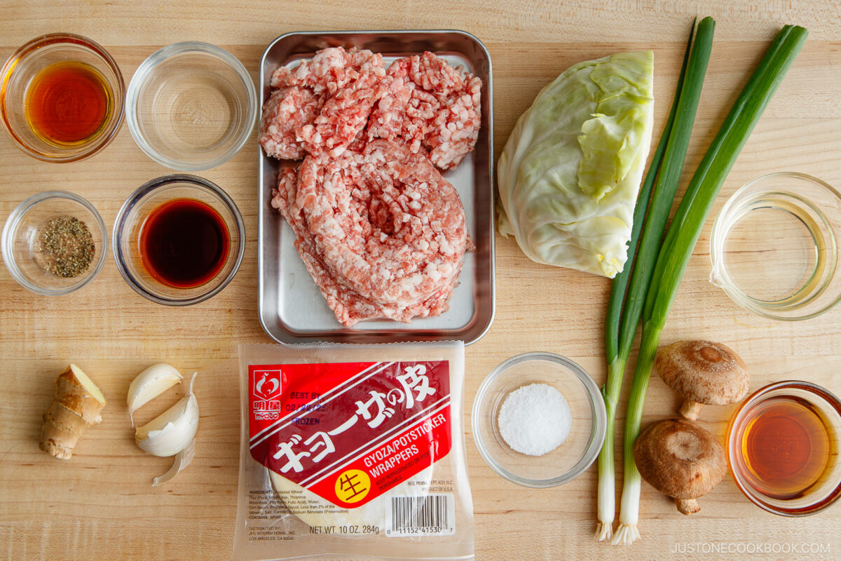 Ingredients for a delicious homemade Gyoza Recipe arranged on a wooden surface: ground meat, gyoza wrappers, cabbage, green onions, shiitake mushrooms, ginger, garlic, seasonings, soy sauce, oil, and vinegar in small glass bowls.