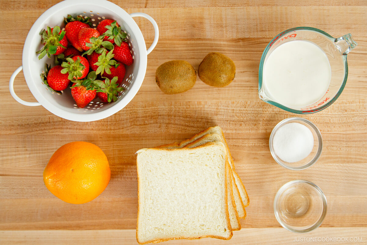 A top-down view of ingredients for a Japanese Fruit Sandwich on a wooden surface: a white colander with strawberries, two kiwis, a measuring cup of milk, an orange, three slices of white bread, sugar, and water.