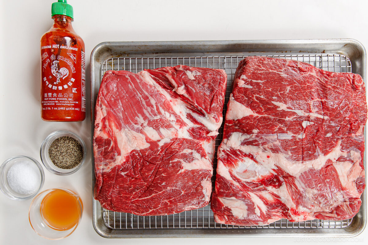 Two large raw beef briskets on a wire rack over a baking sheet, with small bowls of salt, black pepper, apple cider vinegar, and a bottle of Sriracha hot sauce—perfect for pairing with smoked beef ribs.