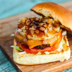 A close-up of a Teriyaki Burger with lettuce, tomato, cheddar cheese, grilled pineapple, crispy onions, and teriyaki sauce on a toasted bun, served on a wooden board.