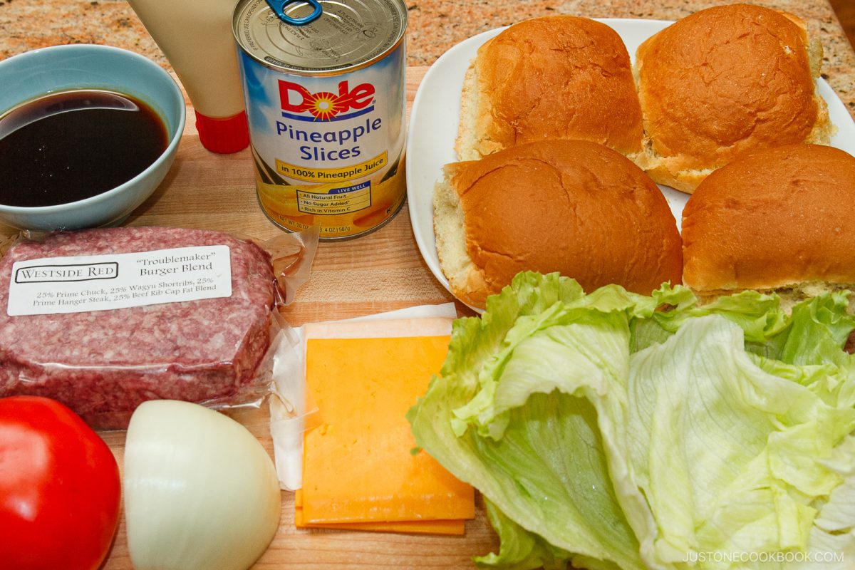 A top view of Teriyaki Burger ingredients, including burger buns, lettuce, cheddar cheese slices, hamburger patties, a tomato, an onion, canned pineapple slices, mayonnaise, and a bowl of dark teriyaki sauce on a countertop.