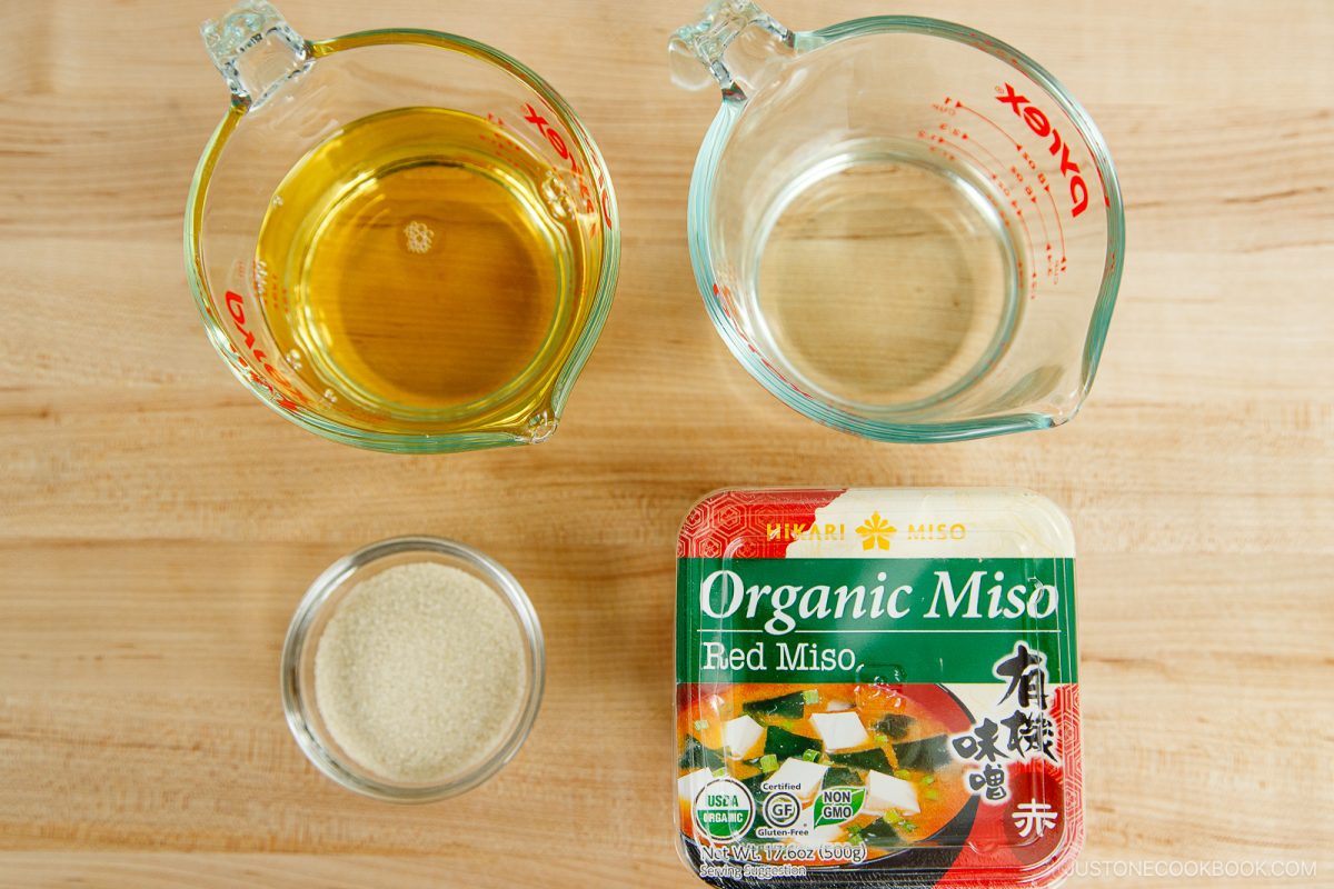 Top-down view of measuring cups with dashi and water, a small bowl of sugar, and a container of organic red miso on a wooden surface—everything you need to create a flavorful All Purpose Miso Sauce.