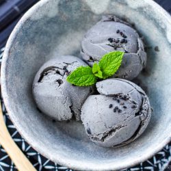 A ceramic bowl with three scoops of rich black sesame ice cream, garnished with a small sprig of mint and sprinkled with black sesame seeds, sits beside a wooden spoon on a patterned cloth.