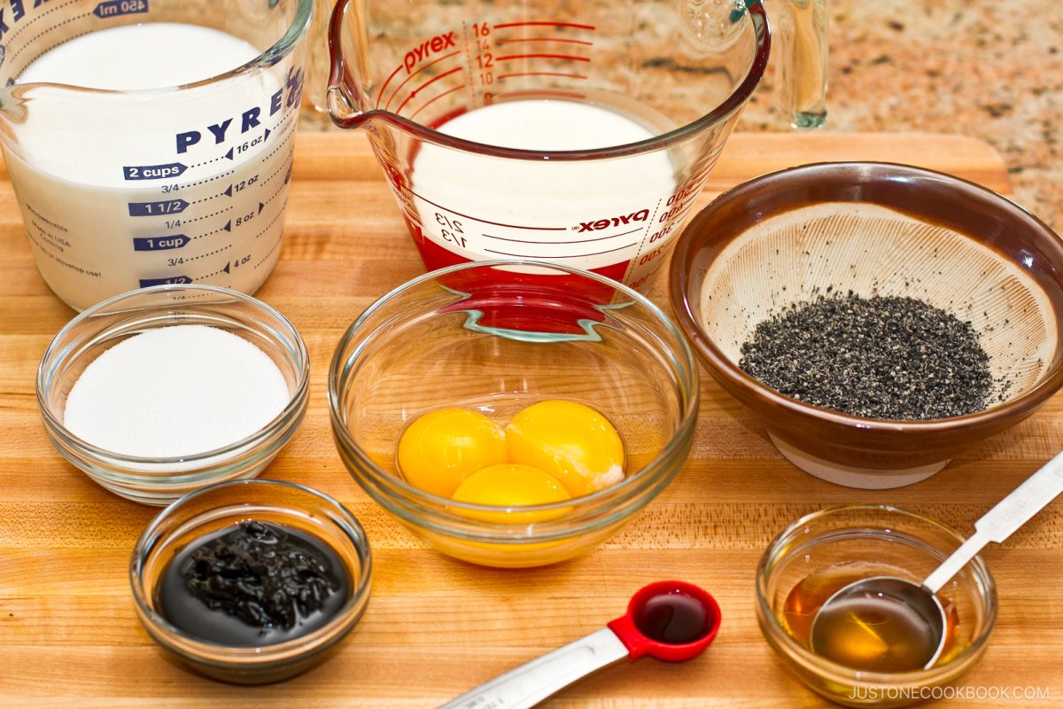 Several bowls on a wooden surface hold egg yolks, black sesame seeds, sugar, milk, cream, black sesame paste, vanilla extract, and a red measuring spoon—ingredients perfectly arranged for making rich and nutty black sesame ice cream.