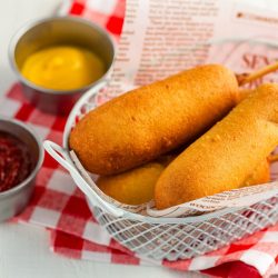 A basket of Japanese corn dogs rests on a red and white checkered cloth, served with small metal cups of ketchup and mustard for dipping. A soda cup is visible in the background.