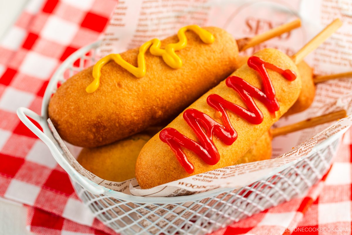 A basket with two Japanese corn dogs on sticks, one topped with yellow mustard and the other with red ketchup, sits on a red and white checkered tablecloth.
