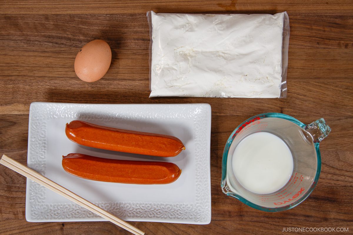 Two hot dogs on a white plate with chopsticks, an egg, a packet of flour, and a measuring cup of milk arranged on a wooden surface—perfect ingredients to make your own Japanese Corn Dog at home.