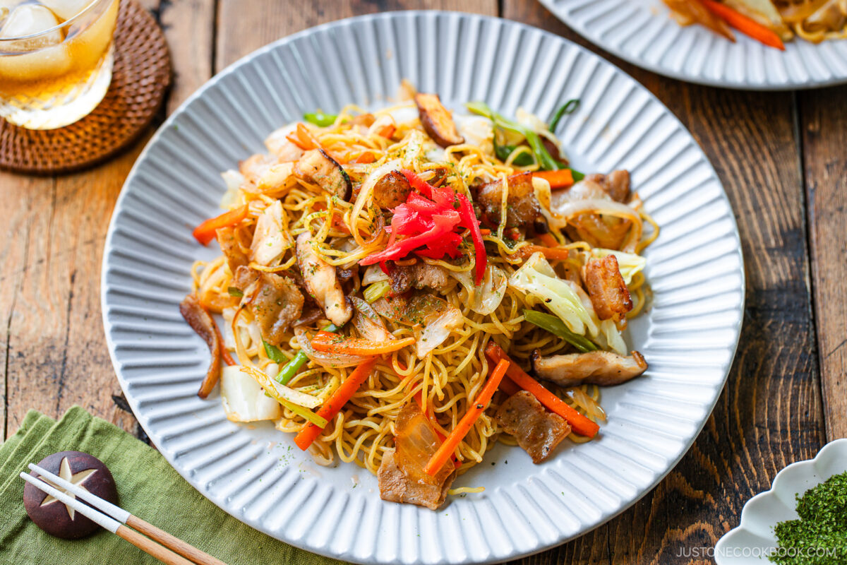 A plate of yakisoba (Japanese stir-fried noodles) topped with sliced pork, vegetables, and pickled ginger, served on a white scalloped plate with chopsticks and a drink on a rustic wooden table.