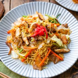 A plate of yakisoba (Japanese stir-fried noodles) topped with sliced pork, vegetables, and pickled ginger, served on a white scalloped plate with chopsticks and a drink on a rustic wooden table.