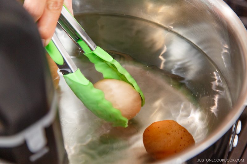 A hand uses green tongs to lift a small potato from a pot of water with other potatoes at the bottom, demonstrating careful handling similar to techniques used in How to Pasteurize Eggs.