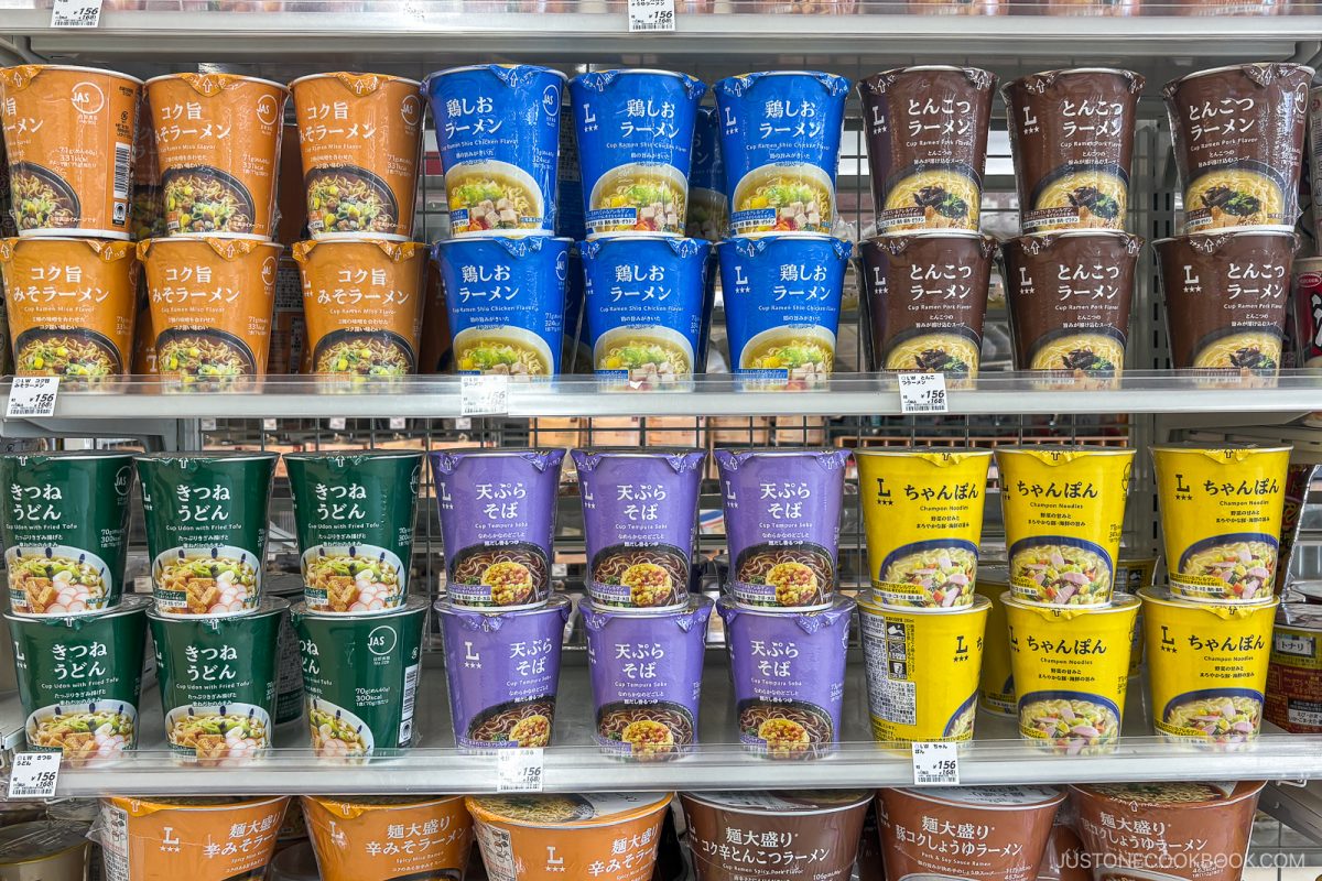 Shelves display colorful instant noodle cups with Japanese labels in orange, blue, brown, green, and yellow, neatly stacked in rows at a grocery store. Price tags are visible under each product—offering shoppers a tempting selection of the best instant ramen in Japan.
