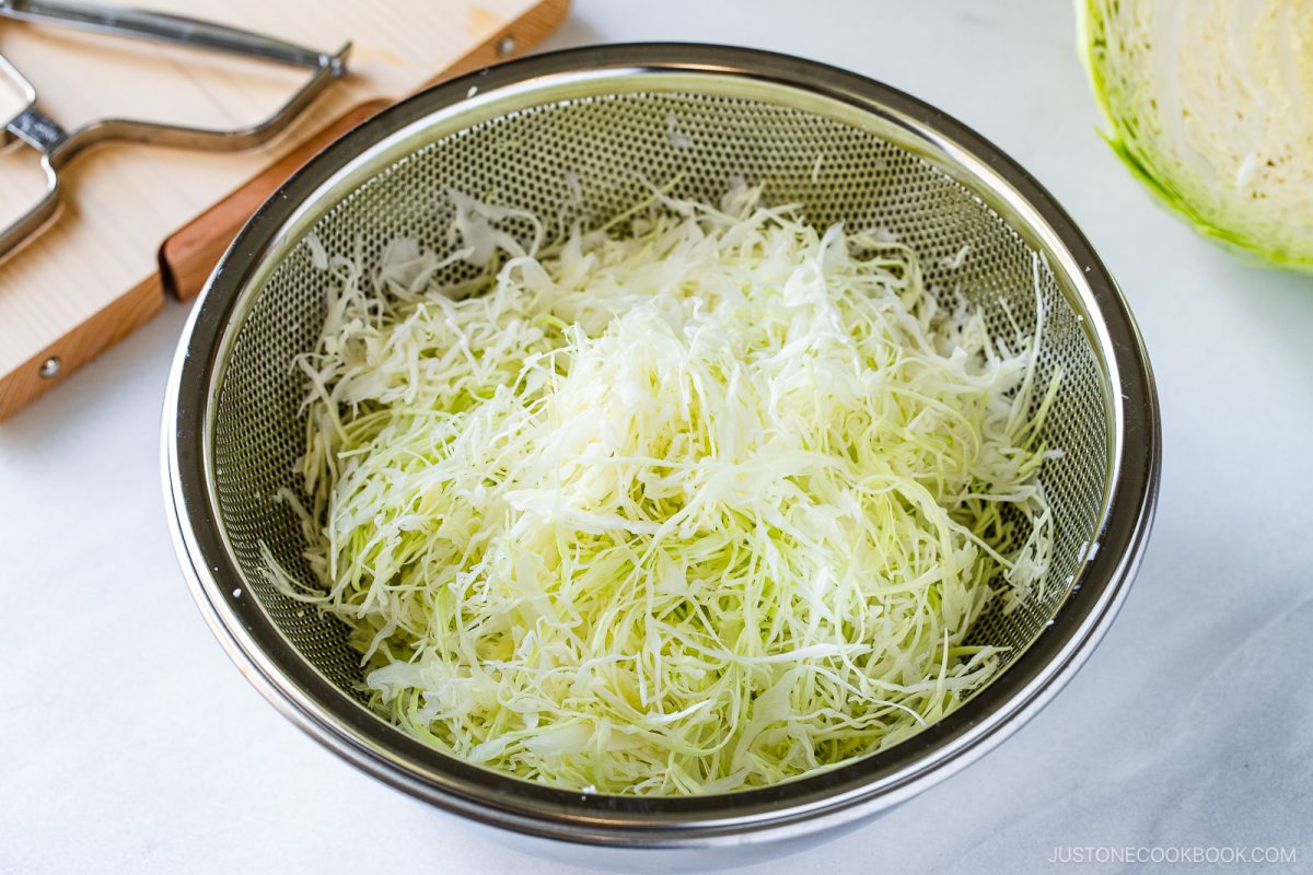 A metal colander filled with finely shredded cabbage, perfect for making a Japanese Shredded Cabbage Salad, sits on a countertop, with a cutting board and kitchen tools in the background.