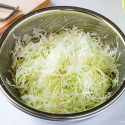 A metal colander filled with finely shredded cabbage, perfect for making a Japanese Shredded Cabbage Salad, sits on a countertop, with a cutting board and kitchen tools in the background.