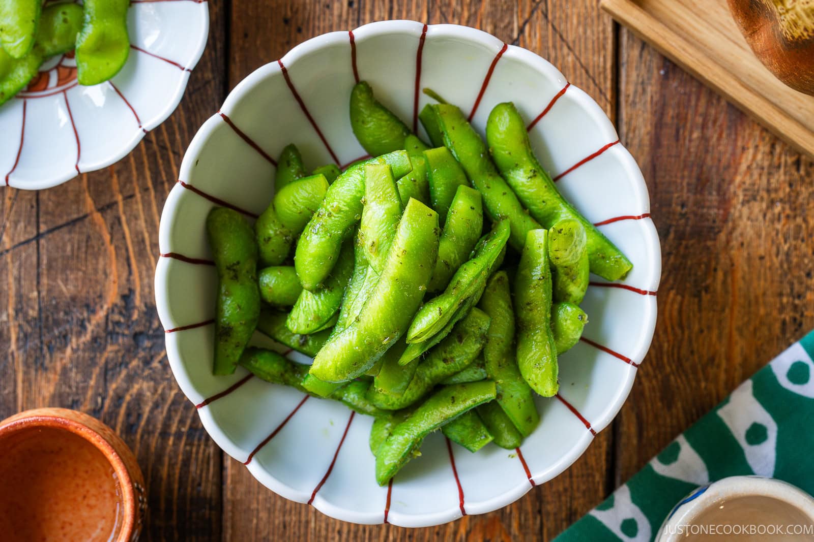 A bowl of bright green yuzu kosho edamame pods sprinkled with salt sits on a wooden table, surrounded by small dishes and a green-and-white patterned cloth.