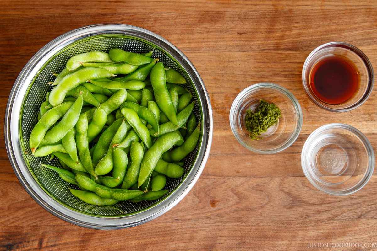 A bowl of fresh Yuzu Kosho Edamame pods in a strainer sits on a wooden table beside three small glass bowls containing soy sauce, green seasoning, and clear water.