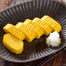 Sliced Dashimaki Tamago (Japanese Dashi Rolled Omelette) is arranged neatly on a dark plate with a small portion of grated daikon radish. The plate rests on a wooden table beside a pair of chopsticks.