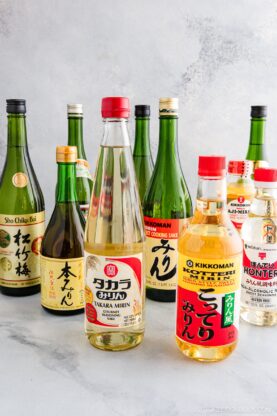 An assortment of Japanese cooking sake, mirin, and rice vinegar bottles arranged on a light gray surface, featuring various brands and colorful labels. Discover Sake vs Mirin: Key Differences and How to Use Them in your kitchen creations.