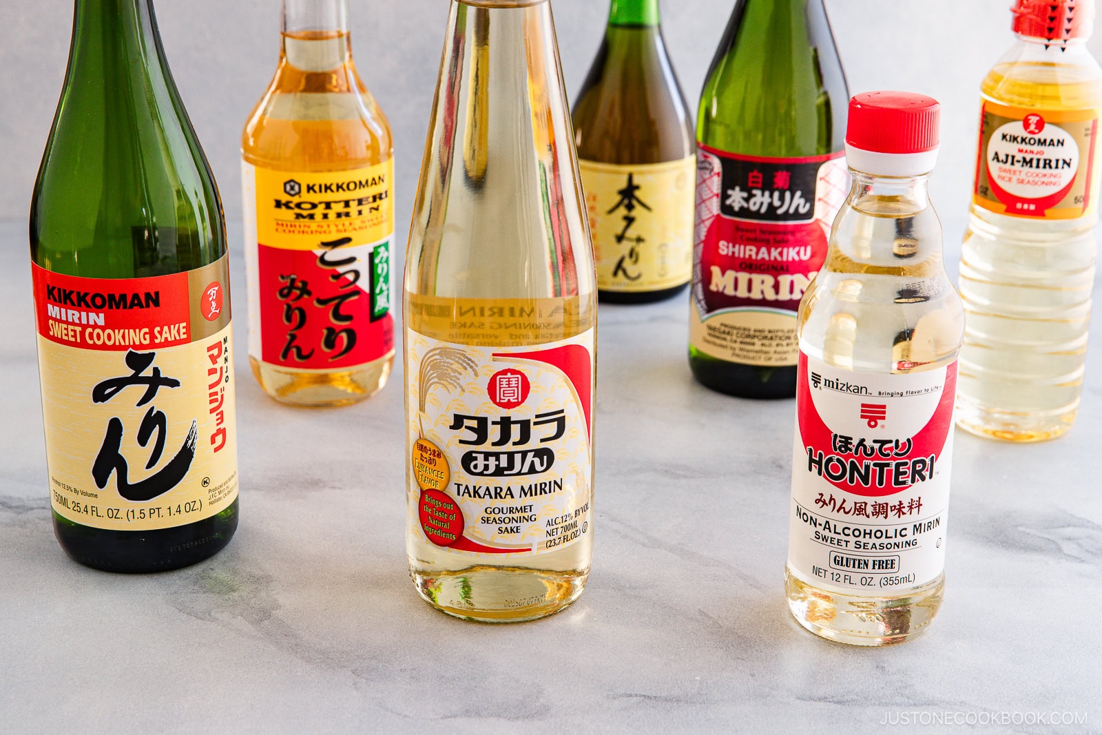 An assortment of Japanese cooking sake and mirin bottles arranged on a light gray surface, featuring various brands and colorful labels in Japanese and English—perfect for exploring Sake vs Mirin: Key Differences and How to Use Them.